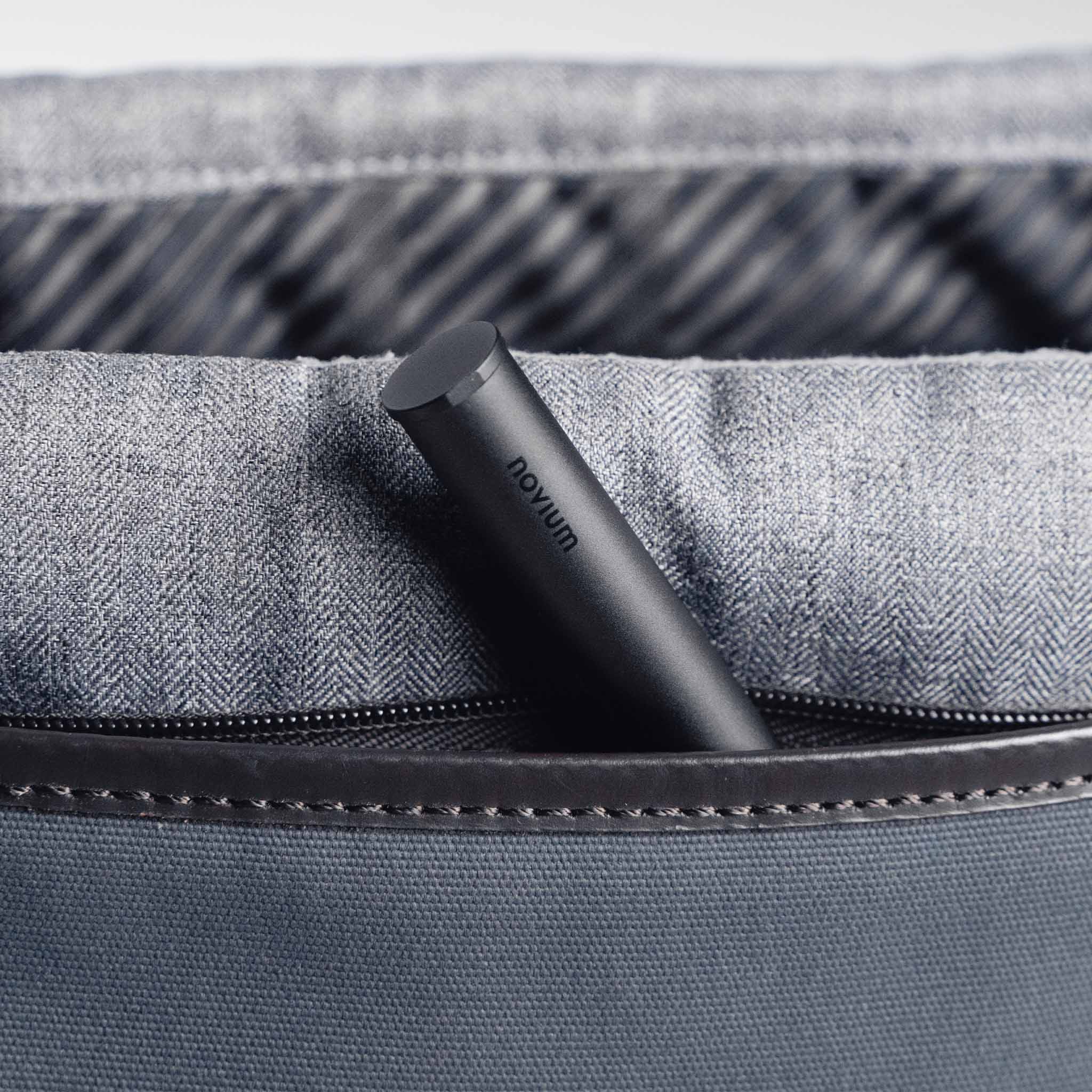 A black aluminum pen case with novum logo is partially visible in a zipped pocket of a grey shoulder bag