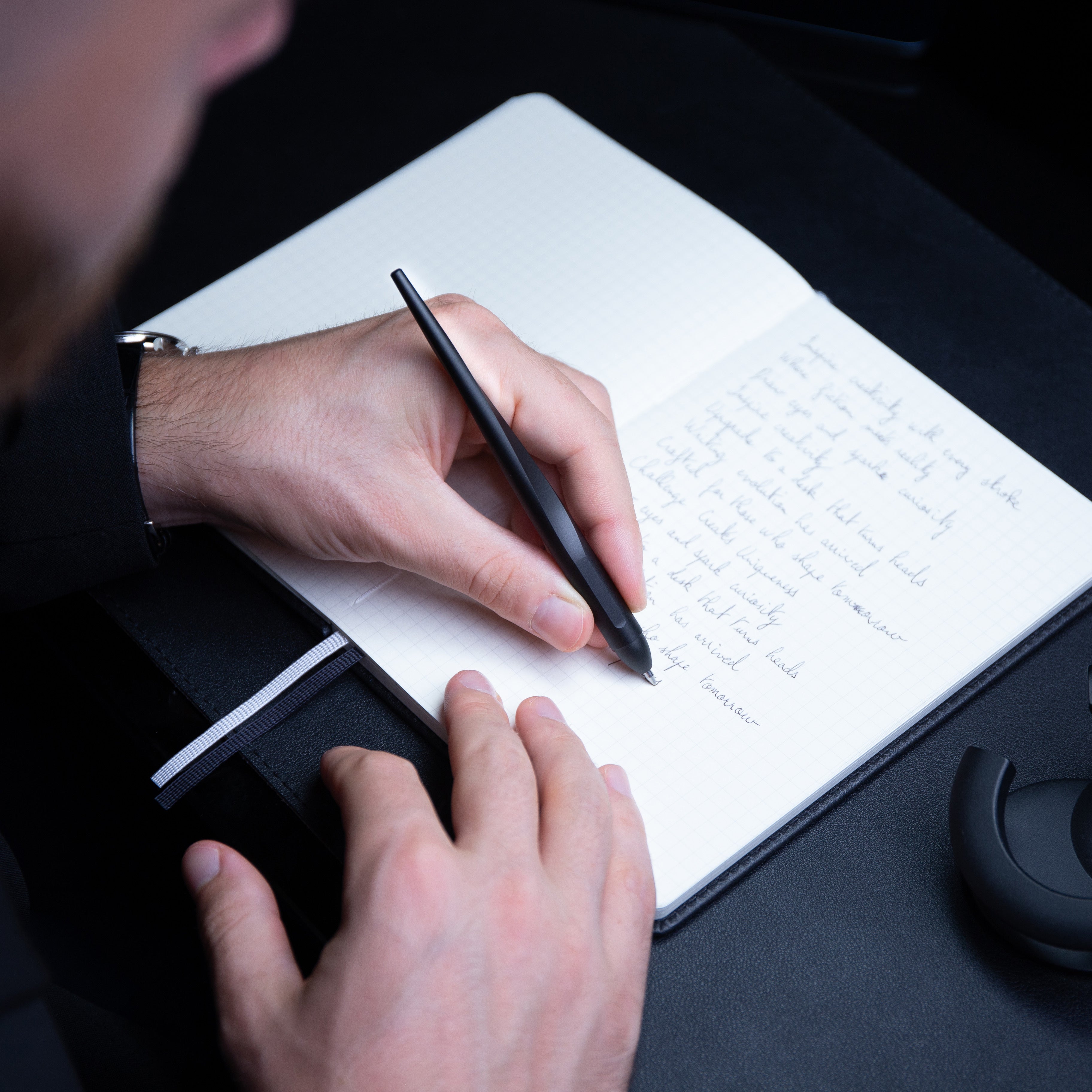 A man writes with a black hoverpen pen in a notebook. The notebook has grided pages and handwritten text